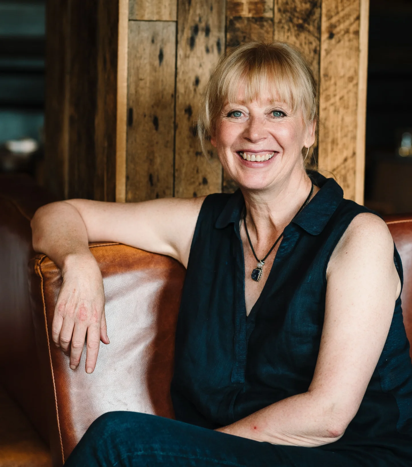 A woman with blonde hair and bangs, wearing a sleeveless dark shirt and necklace, sits smiling on a brown leather couch in front of a wooden wall, radiating plentiful wellbeing and positivity.