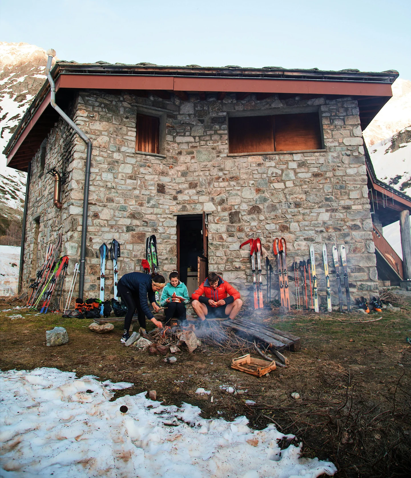 Three people sit and crouch around a small campfire outside a stone mountain cabin, surrounded by snow and several pairs of skis propped against the building.