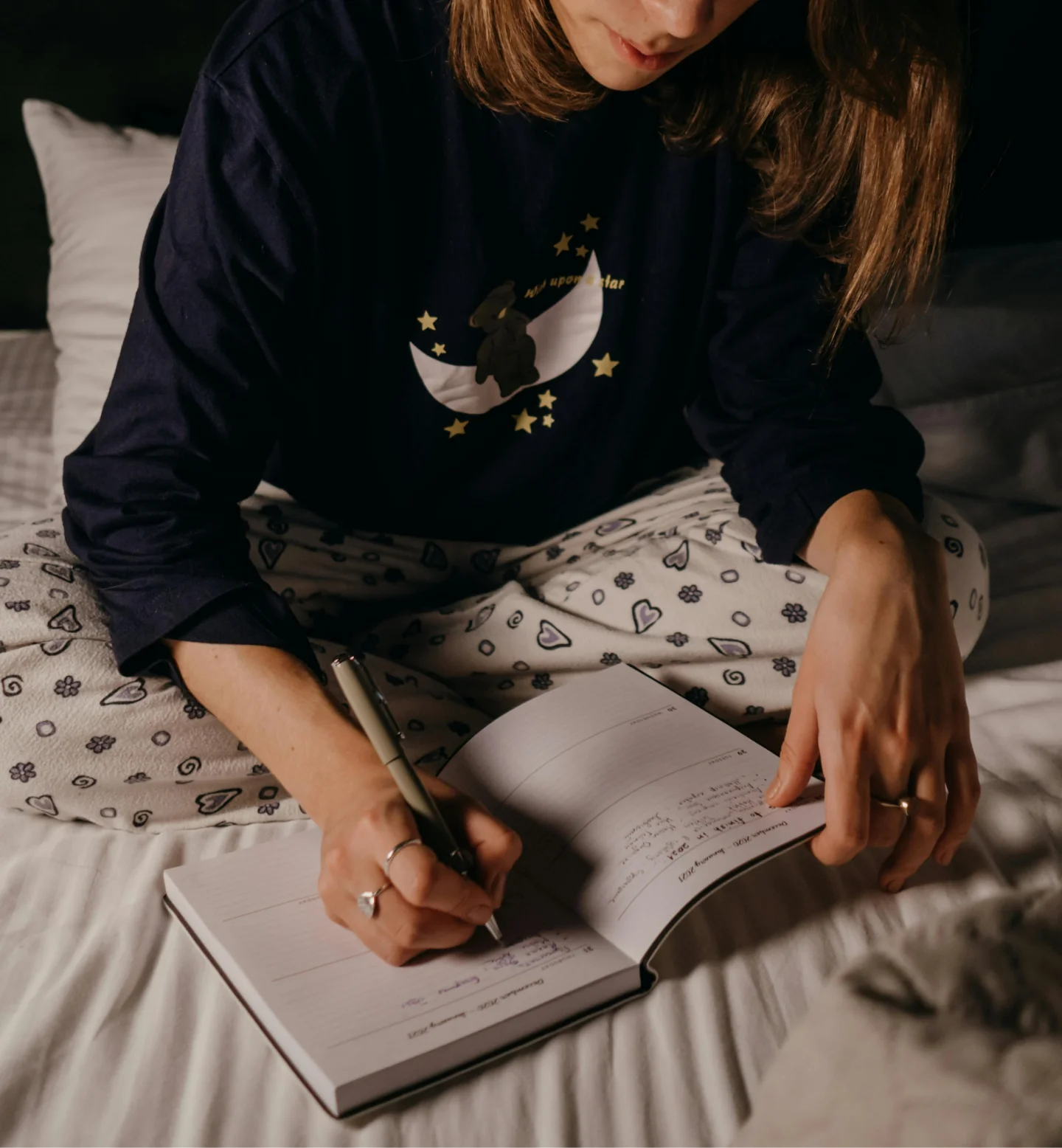 A person sitting cross-legged on a bed in pajamas writes in a journal or notebook. The pajamas have heart patterns, and the top features a moon and stars design. The photo is taken indoors in a cozy, dimly lit setting.