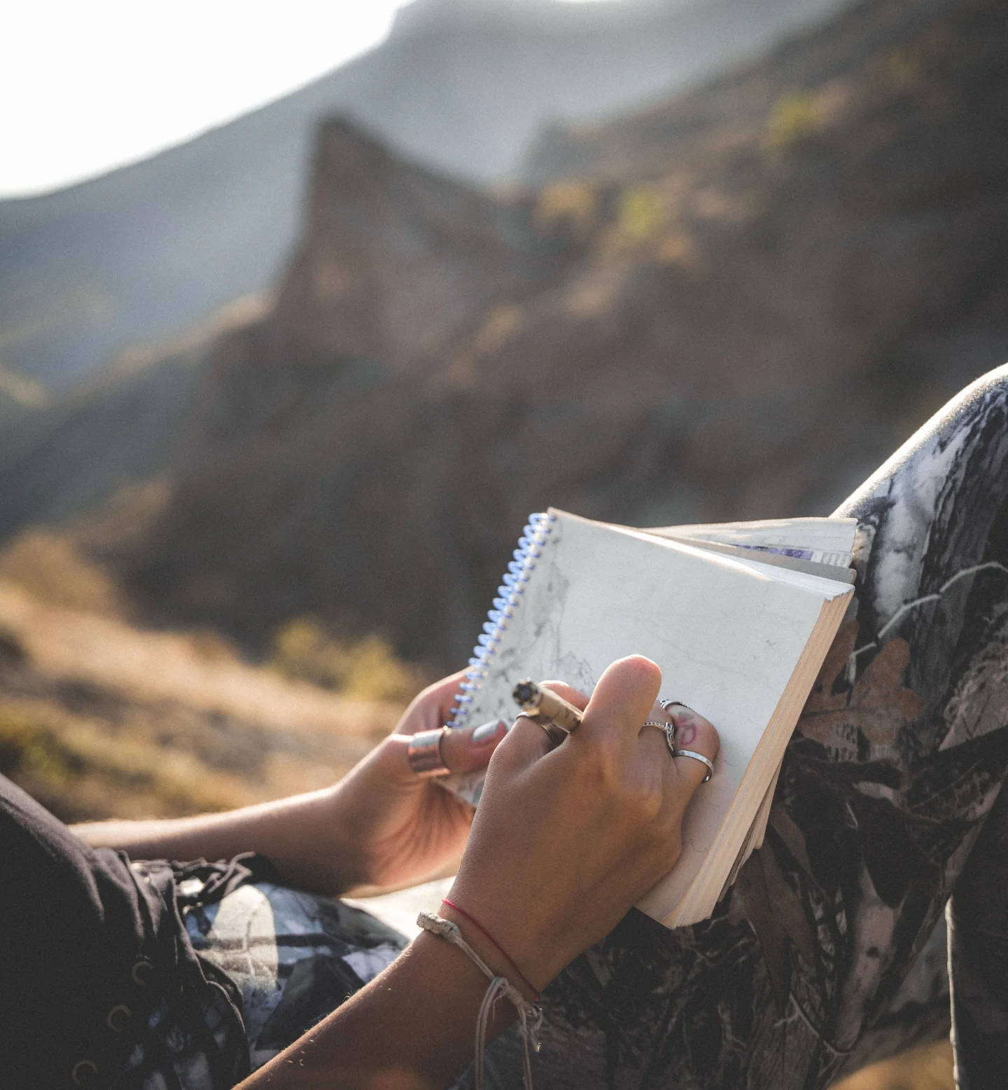 A person wearing patterned pants sits outdoors in a mountainous area, holding a pen and sketching or writing in a spiral notebook on their lap. Sunlight gently illuminates the scene.