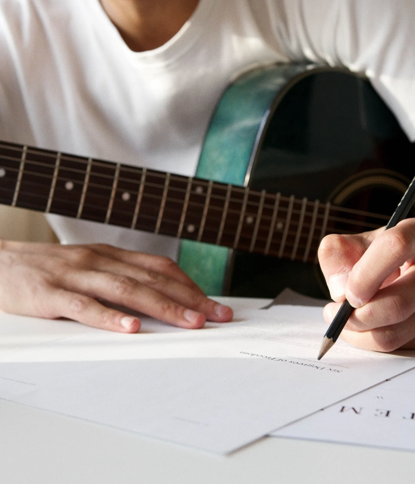 A person wearing a white shirt holds a guitar and writes on sheets of paper with a pen, suggesting they are composing music or writing lyrics.