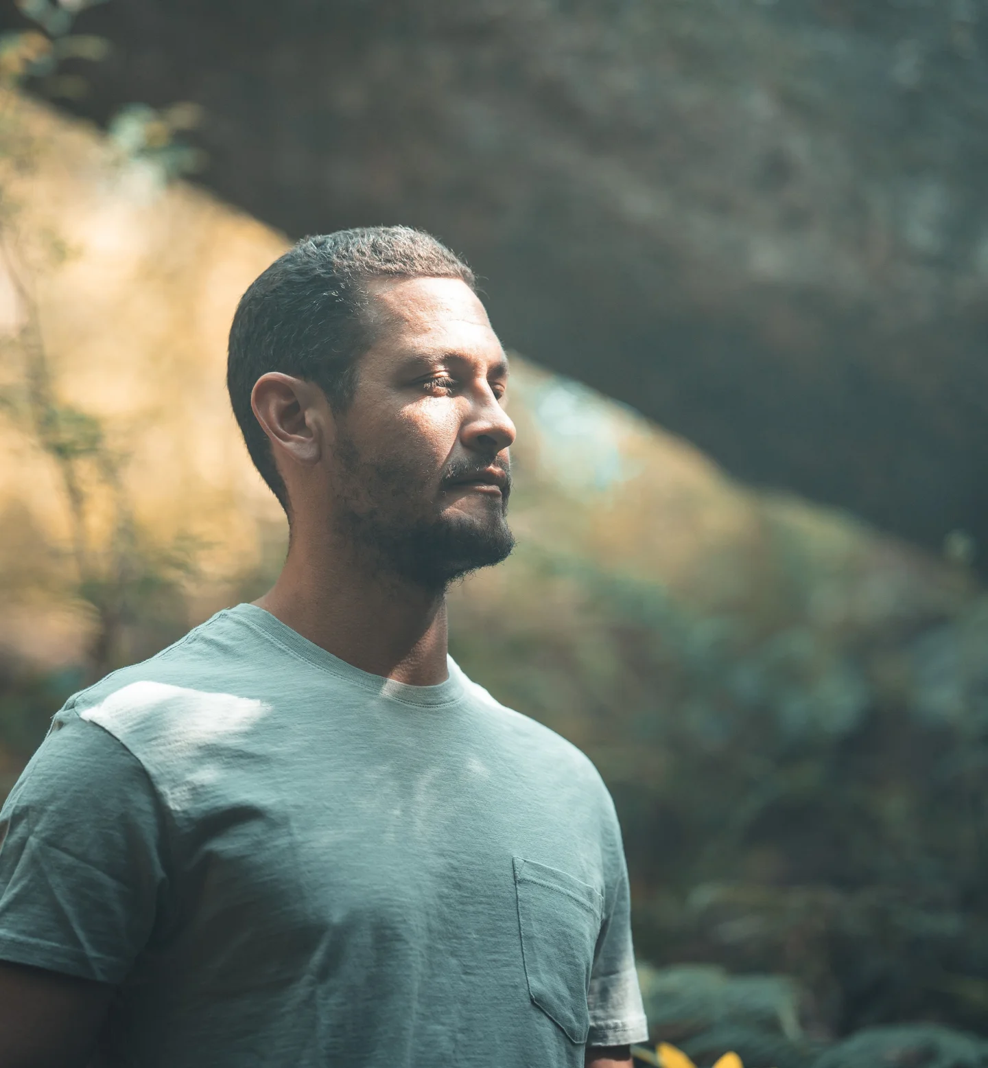 A man with short hair and a beard stands outdoors with his eyes closed, wearing a light gray t-shirt. Sunlight filters through trees, creating a peaceful, natural background.