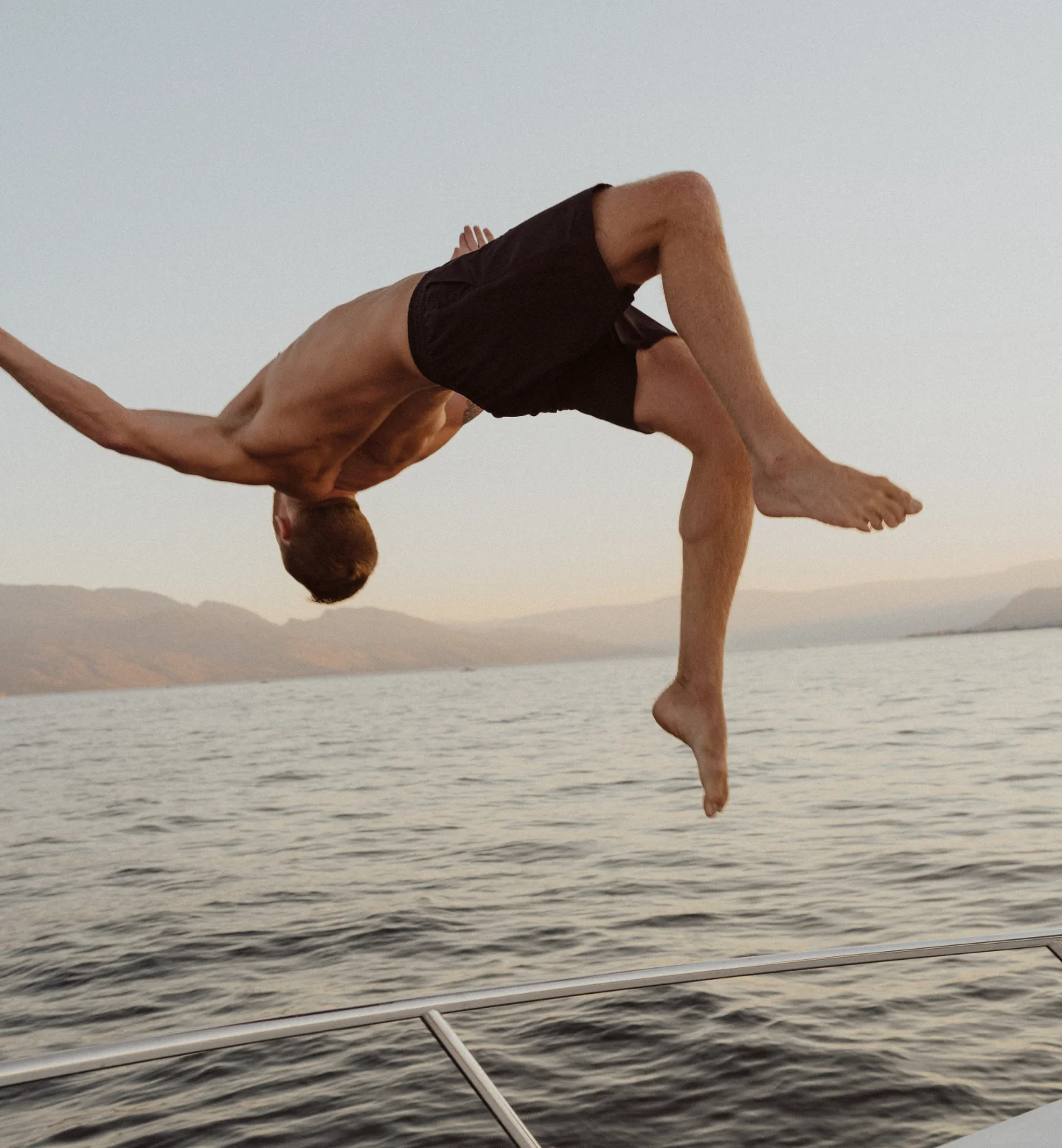 A shirtless person in black shorts is mid-backflip off the edge of a boat, with a calm body of water and distant mountains visible under a clear sky at sunset.