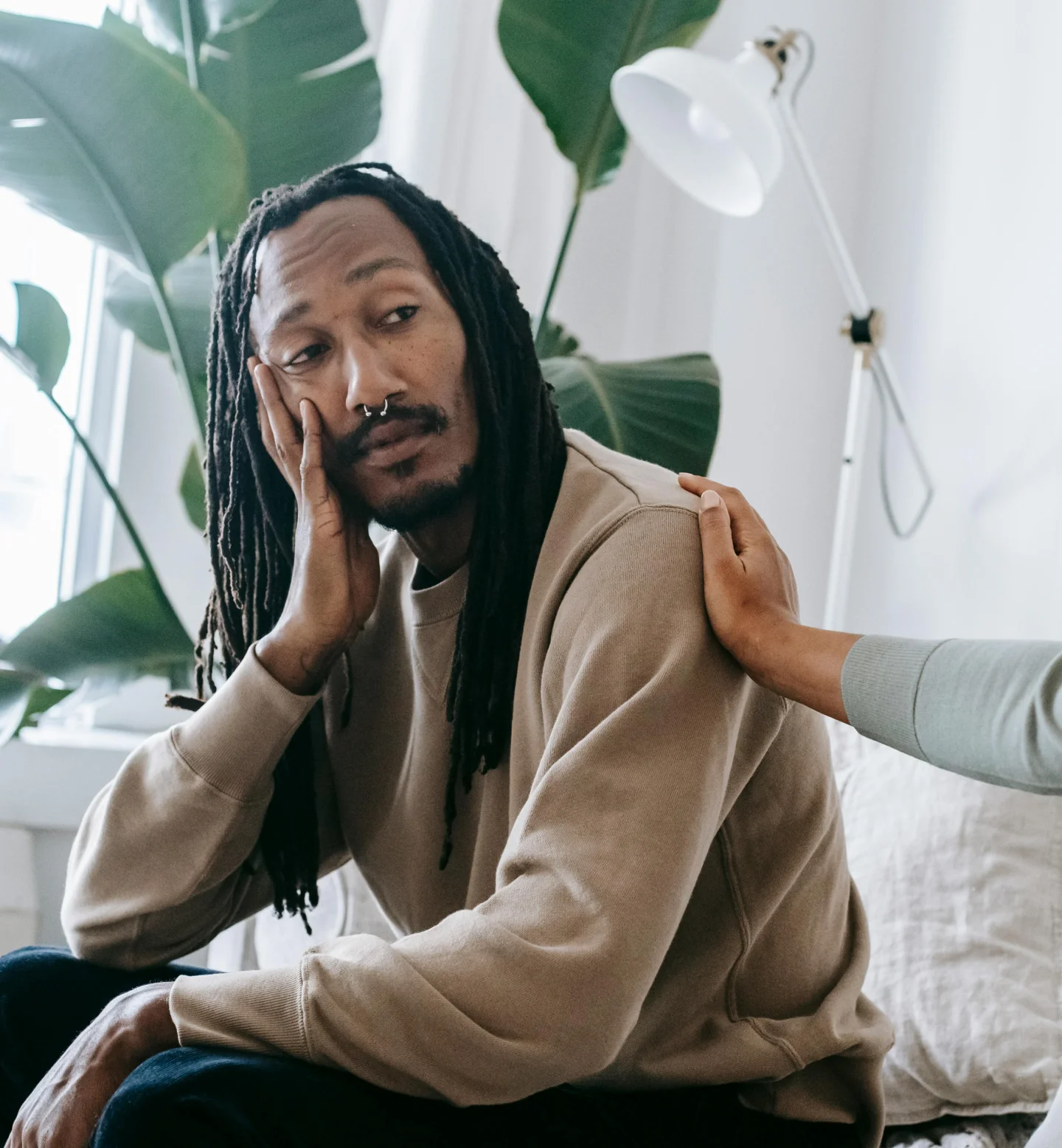 A person with long dreadlocks and a nose ring sits on a couch, resting their face on one hand and looking thoughtful, while another person gently places a hand on their shoulder. Large green plants are in the background.
