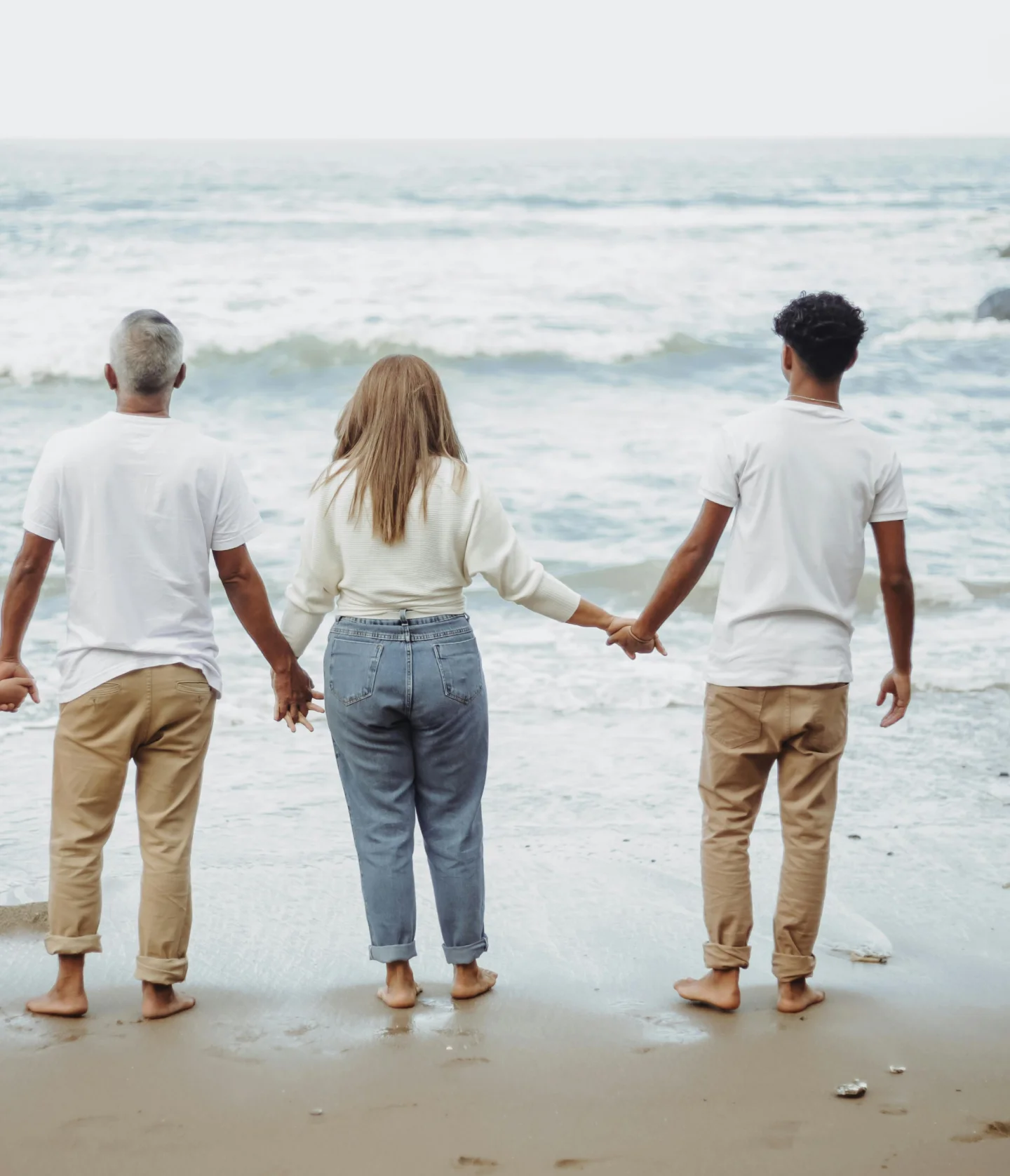 Three people stand barefoot on the beach, holding hands and facing the ocean. They are dressed in casual clothes and appear to be enjoying a peaceful moment together by the water.