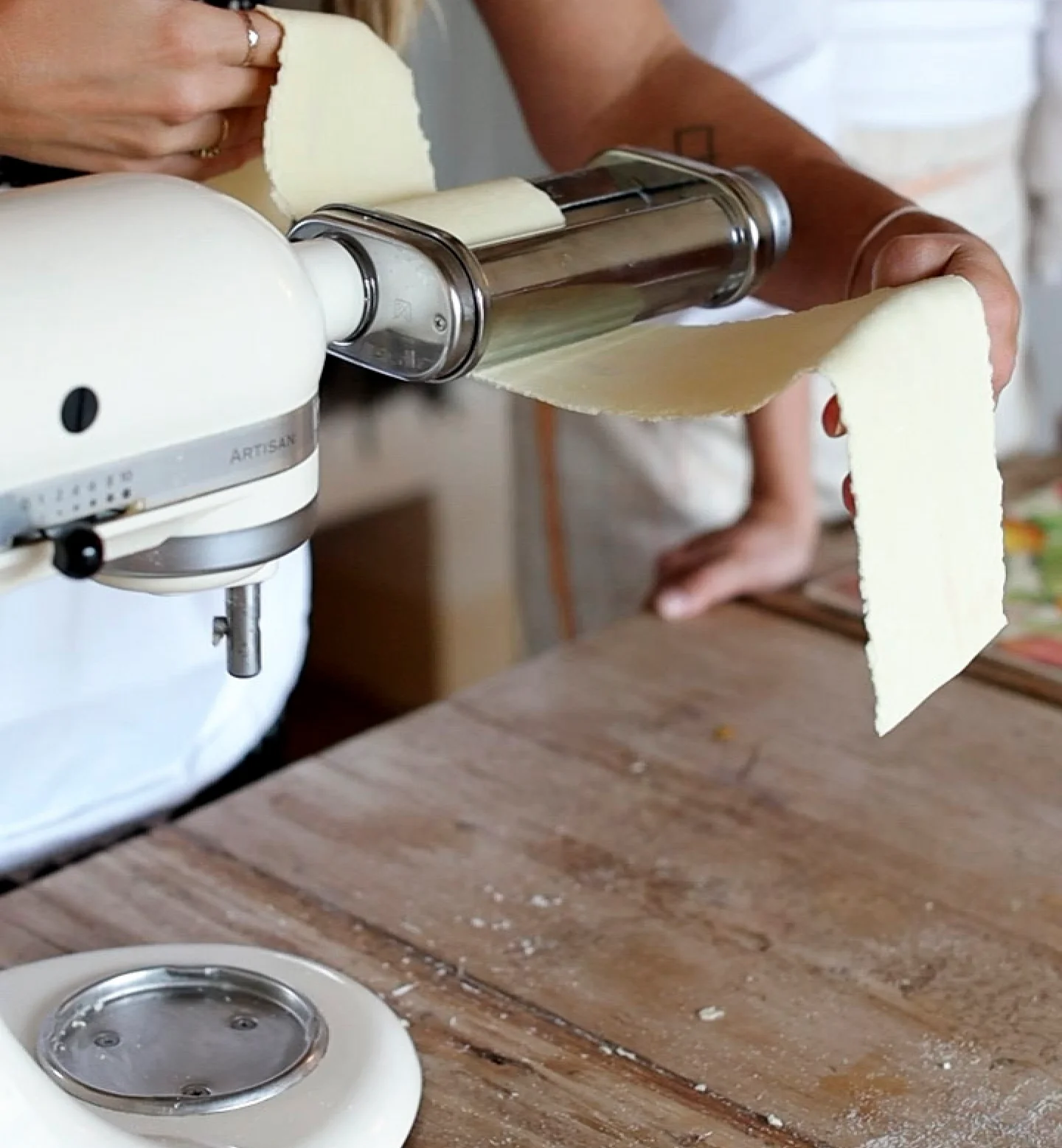 A person feeds a sheet of dough through a pasta roller attachment on a stand mixer, making fresh pasta on a wooden table.