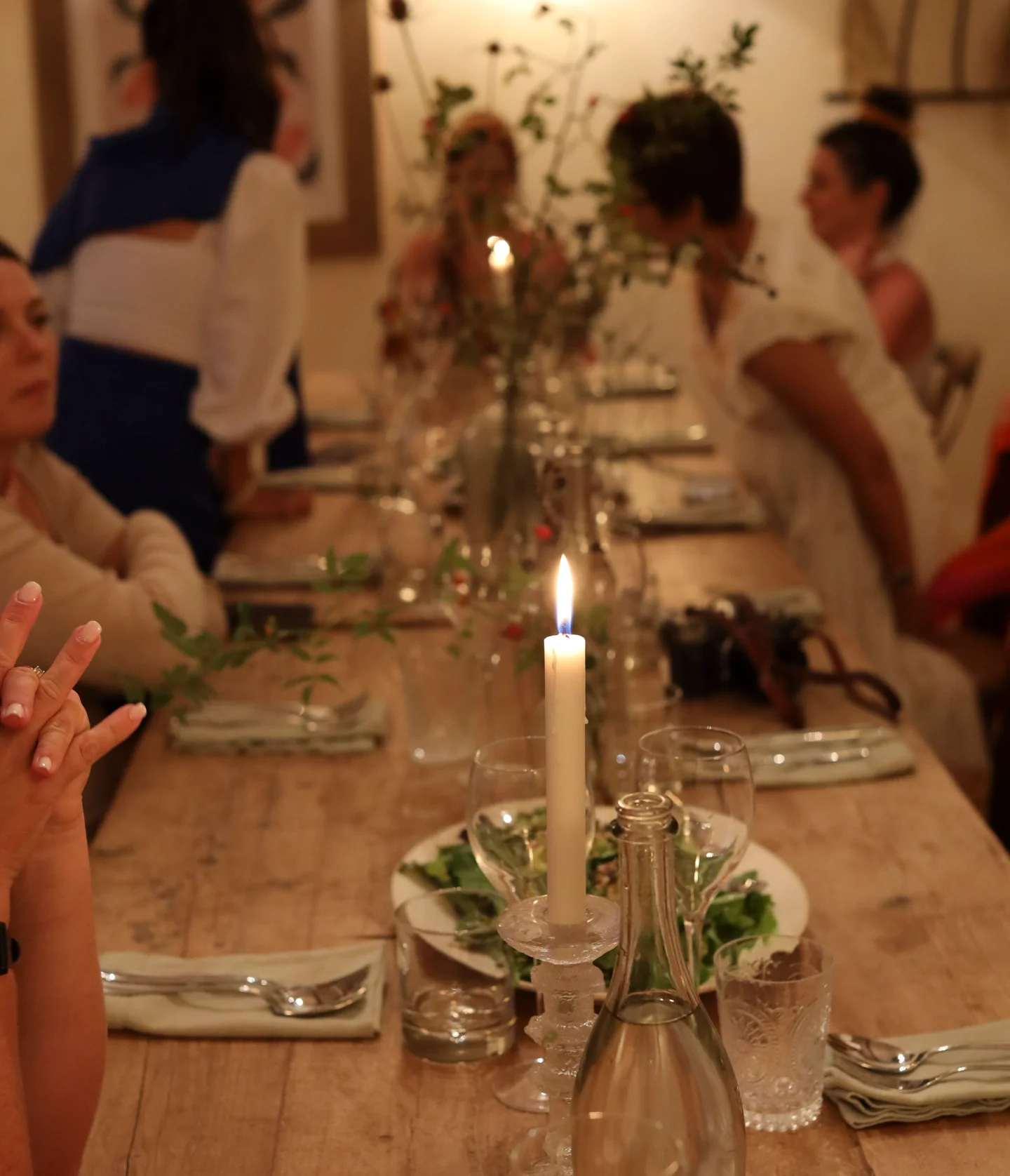 A lit candle stands on a rustic wooden table set for dinner, surrounded by glasses, plates, and greenery, while several people sit and chat in a softly lit, cozy room.
