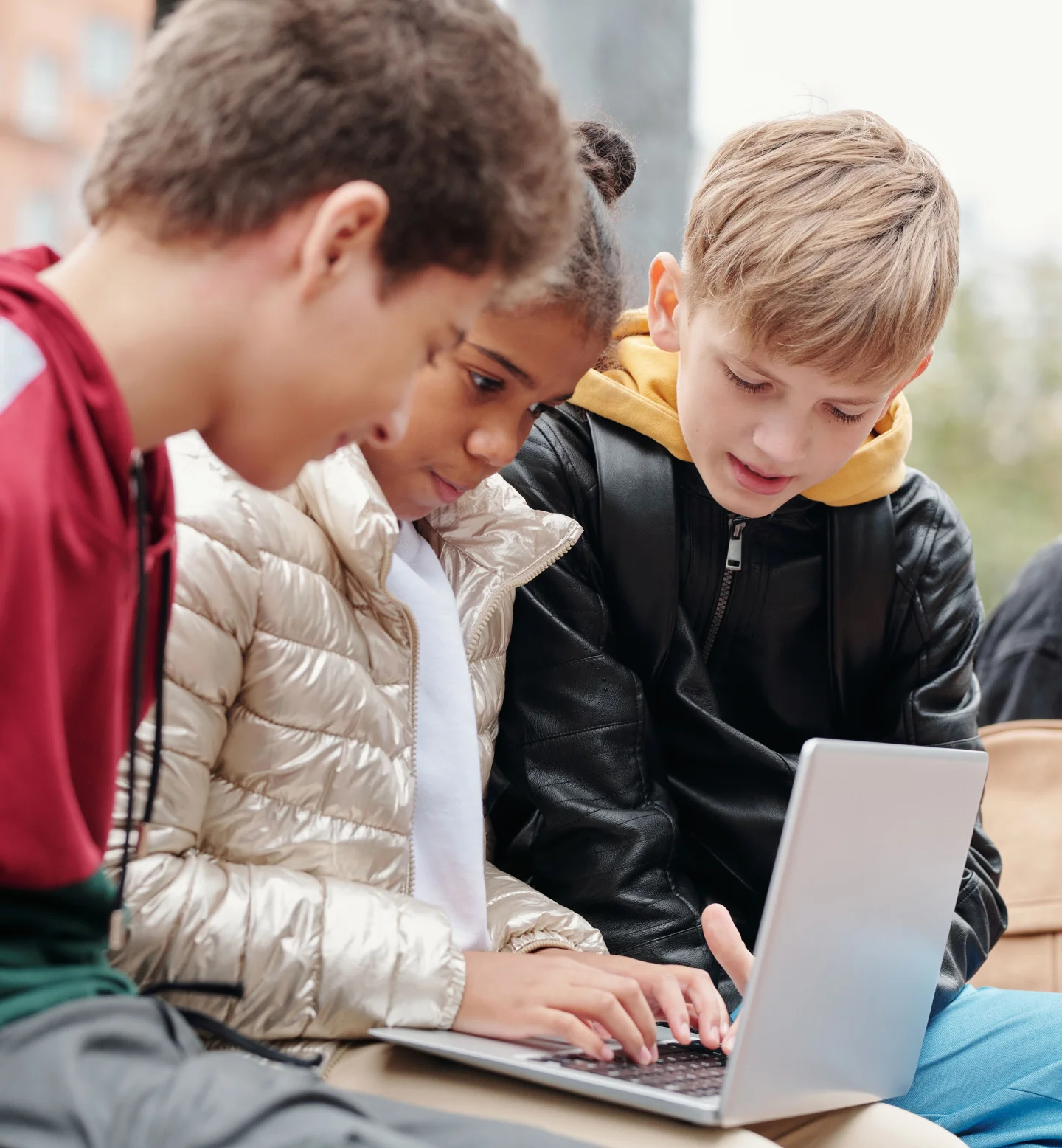 Three children sit closely together outdoors, looking at and using a laptop. They appear to be collaborating or sharing something on the screen, with focused and engaged expressions.