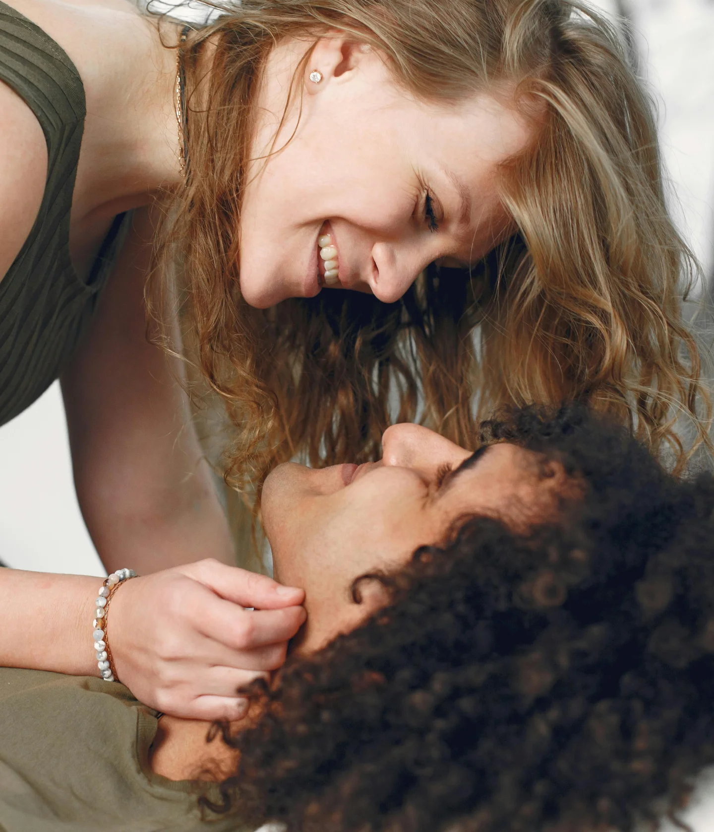 A smiling woman with long blonde hair leans over a man with curly hair, gently cupping his face as they gaze at each other lovingly, sharing a joyful and intimate moment.