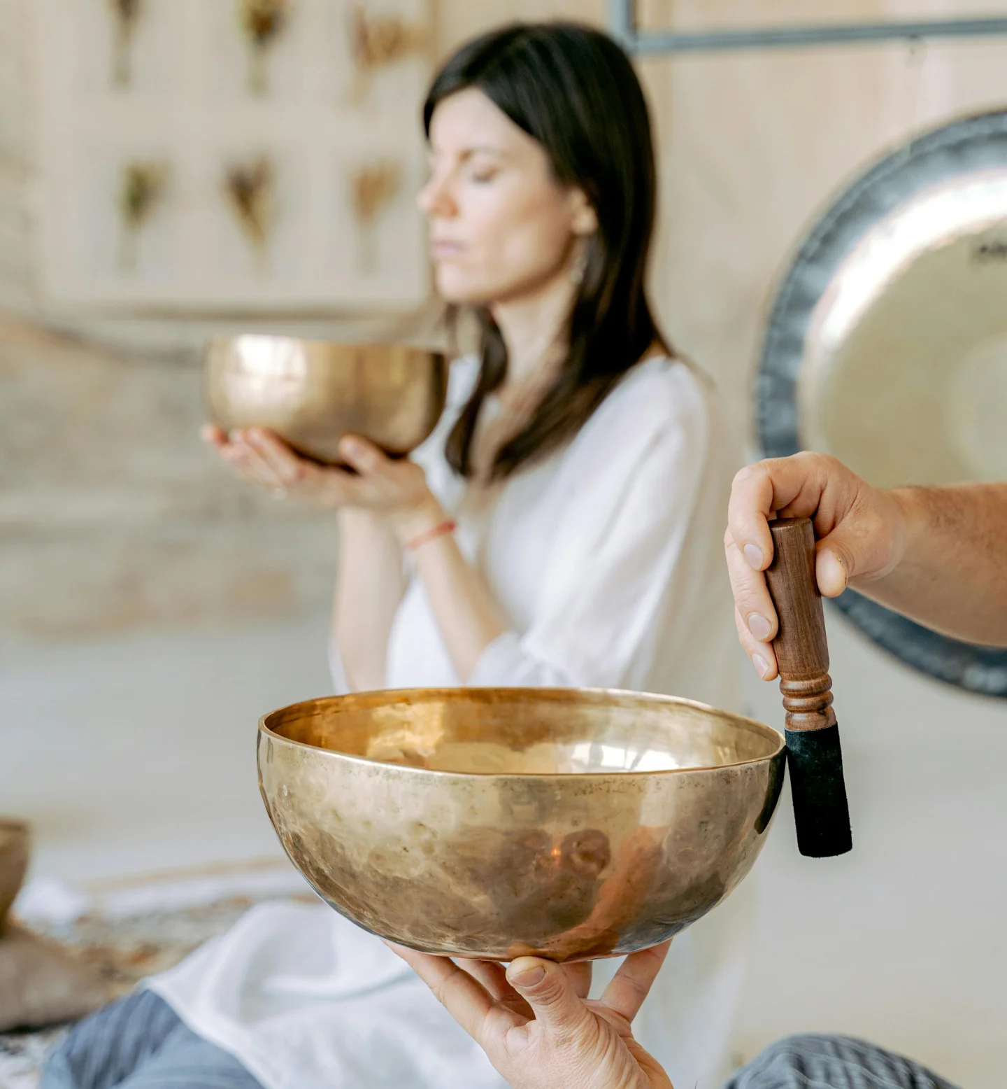 A woman in white holds a brass singing bowl with closed eyes, meditating. In the foreground, another hand holds a singing bowl and a mallet. A large gong is seen in the background.