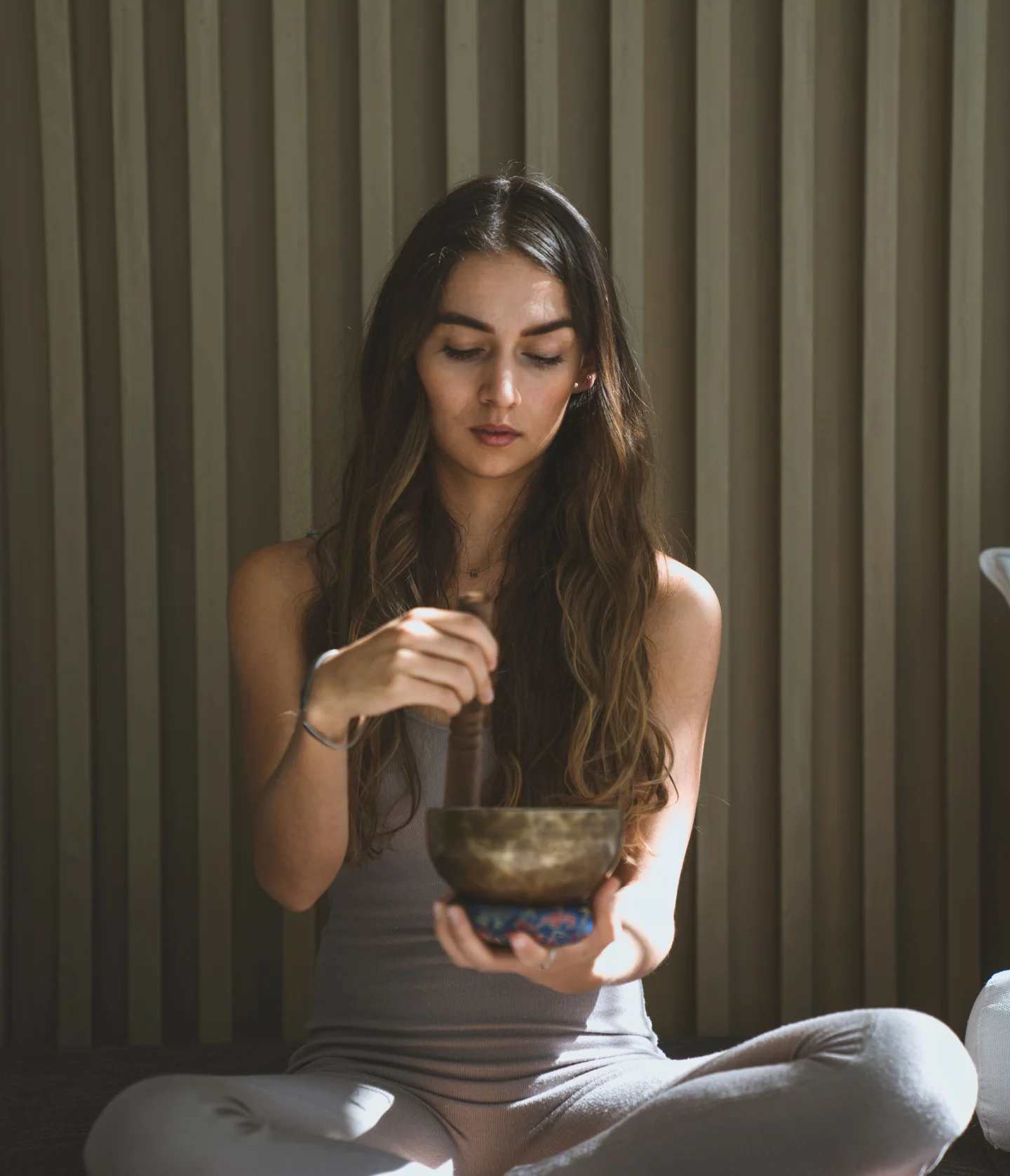 A woman with long brown hair sits cross-legged, wearing light-colored clothes, and holds a singing bowl, gently striking it with a mallet. She appears calm and focused, with a striped wall behind her.