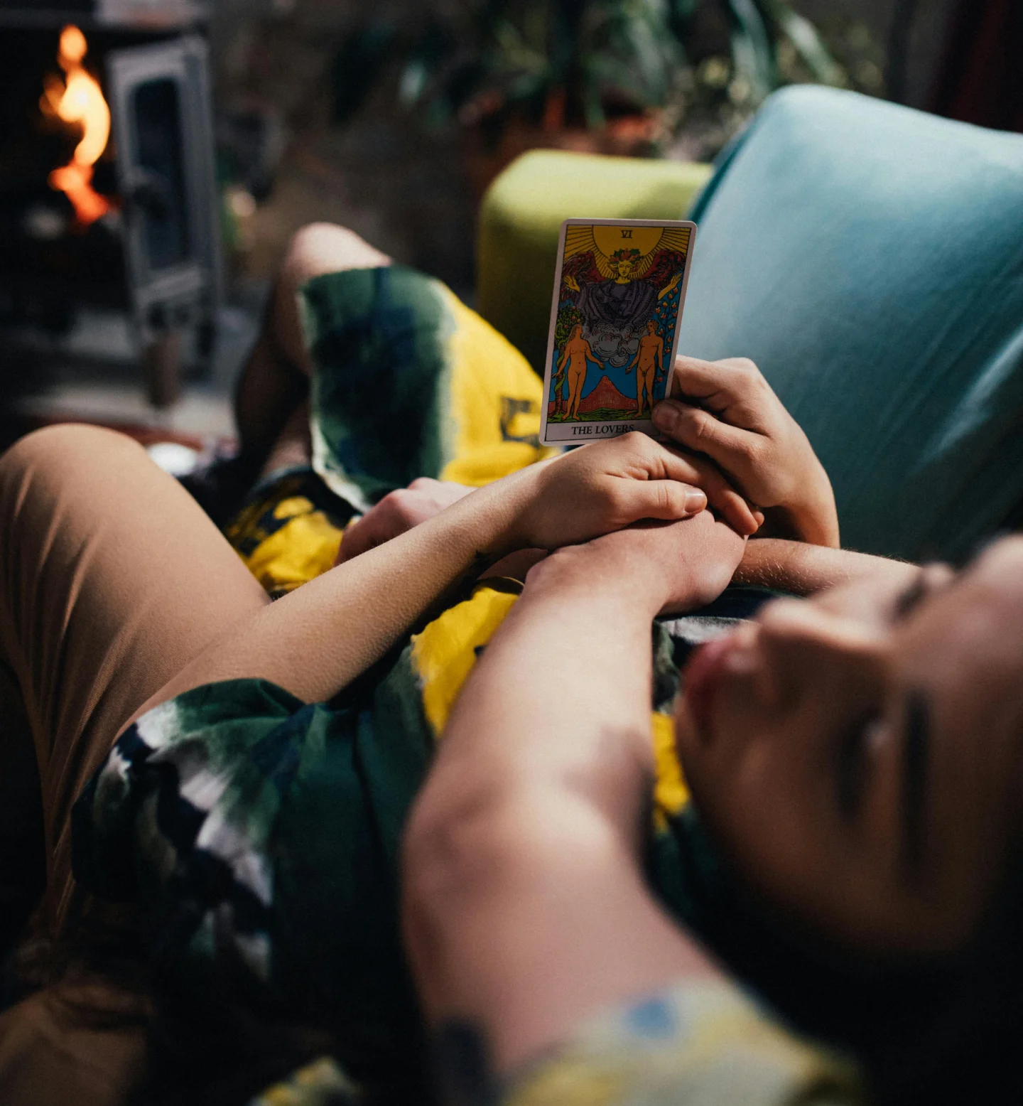 A person lying on a couch holds up The Lovers tarot card while another person rests nearby; a fireplace and plants are visible in the cozy background.