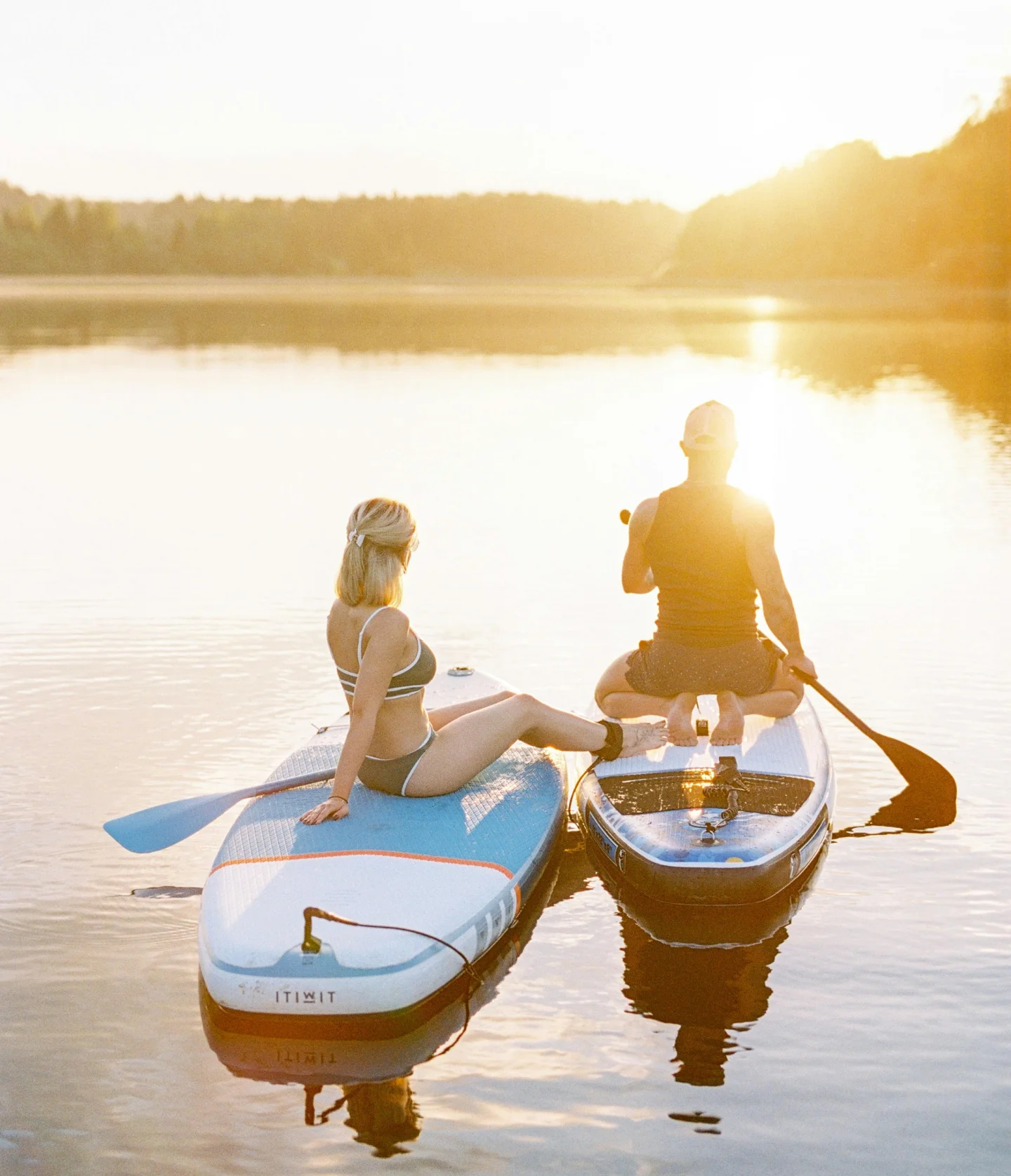 Two people on paddleboards sit and relax on a calm lake during a golden sunset, surrounded by gentle water and distant tree-lined shores.