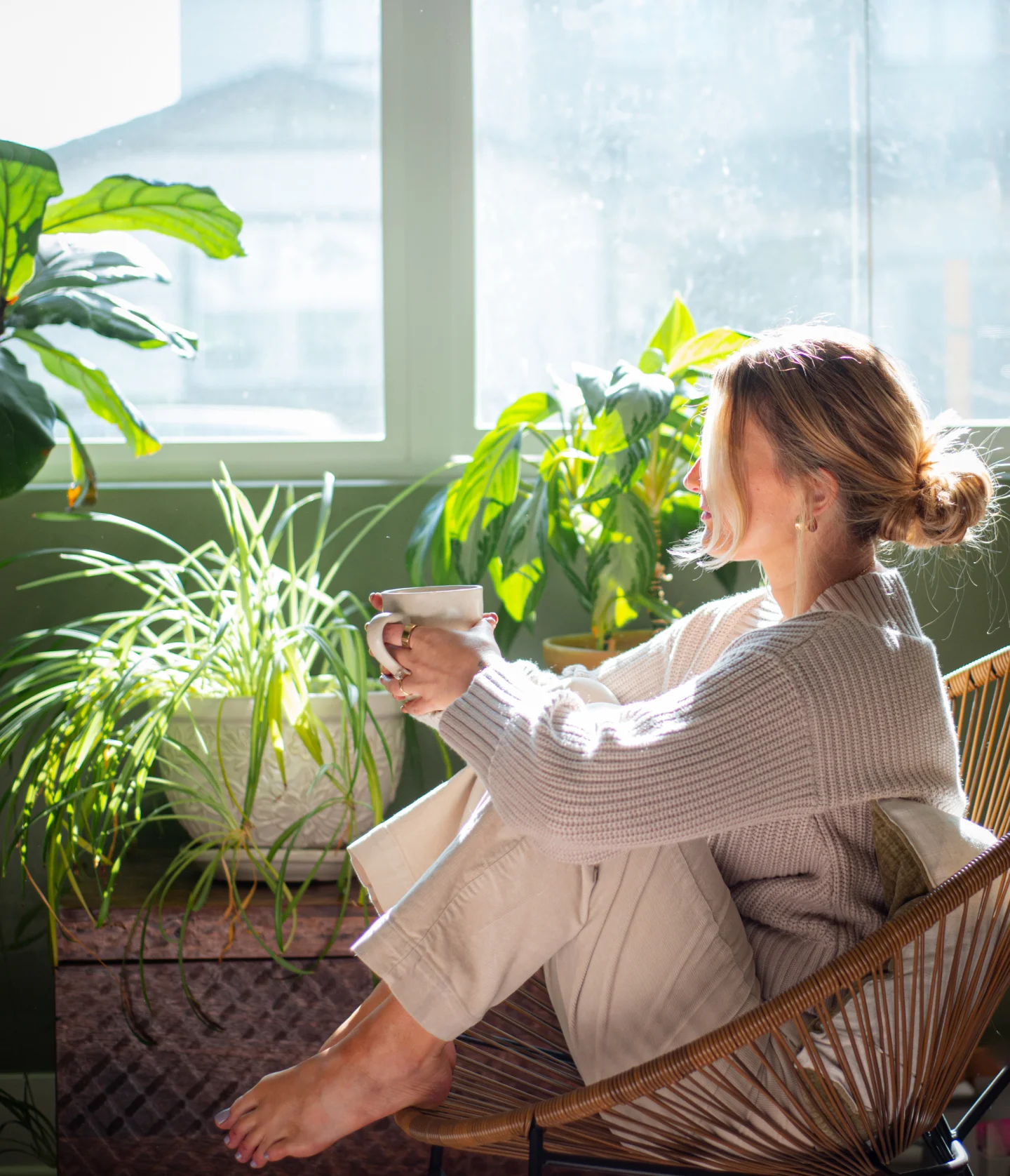 A woman sits barefoot in a chair by a sunlit window, holding a mug and surrounded by green houseplants. She is wearing light-colored, cozy clothes and gazes outside, bathed in natural light.
