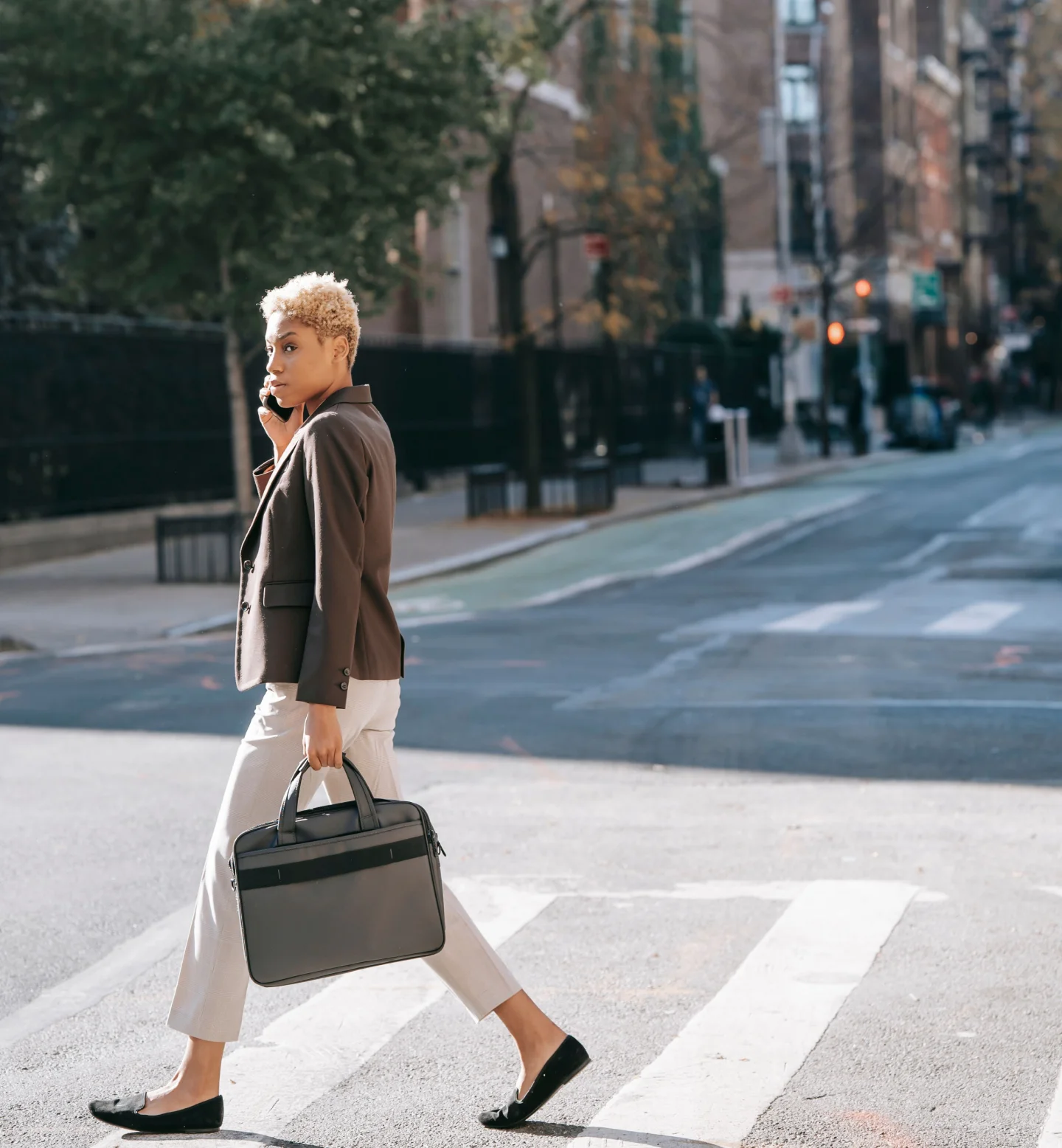 A person with short blond hair, wearing a brown blazer and white pants, crosses a city street on a crosswalk while talking on a phone and carrying a large black handbag. Trees and buildings line the street in the background.