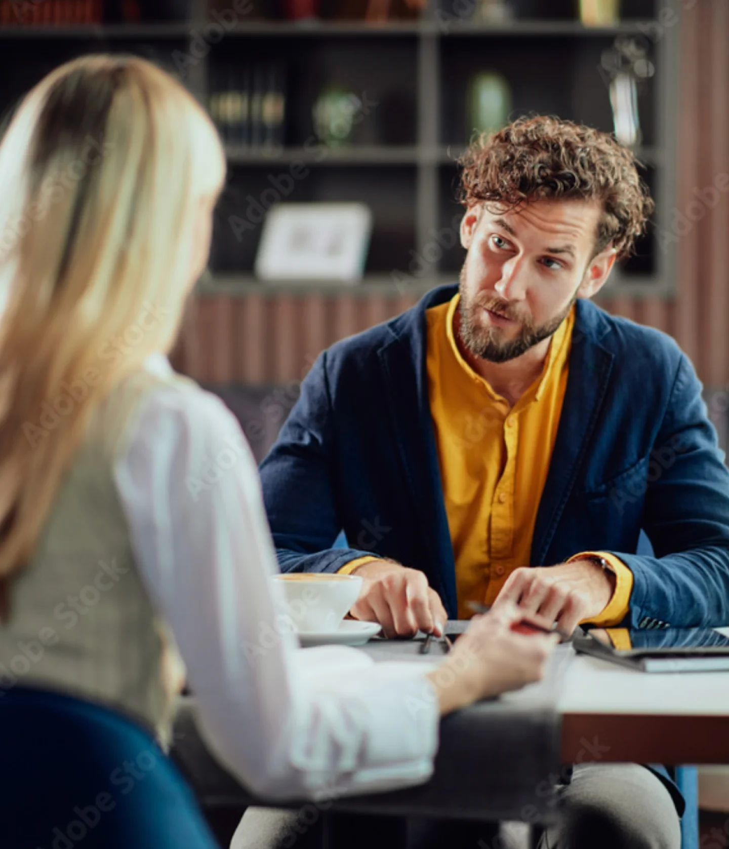 A man with curly hair and a beard, wearing a yellow shirt and blue jacket, sits at a table talking to a woman with long blonde hair in an office or library setting. They appear to be having a serious conversation.