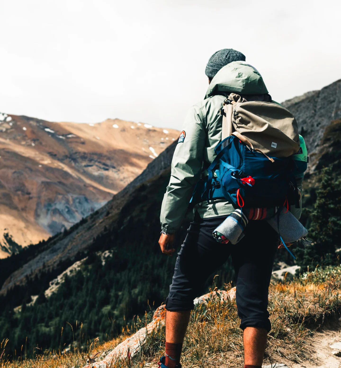 A person wearing outdoor gear and a backpack stands on a grassy slope, overlooking a mountainous landscape with patches of snow and forested valleys under a cloudy sky.