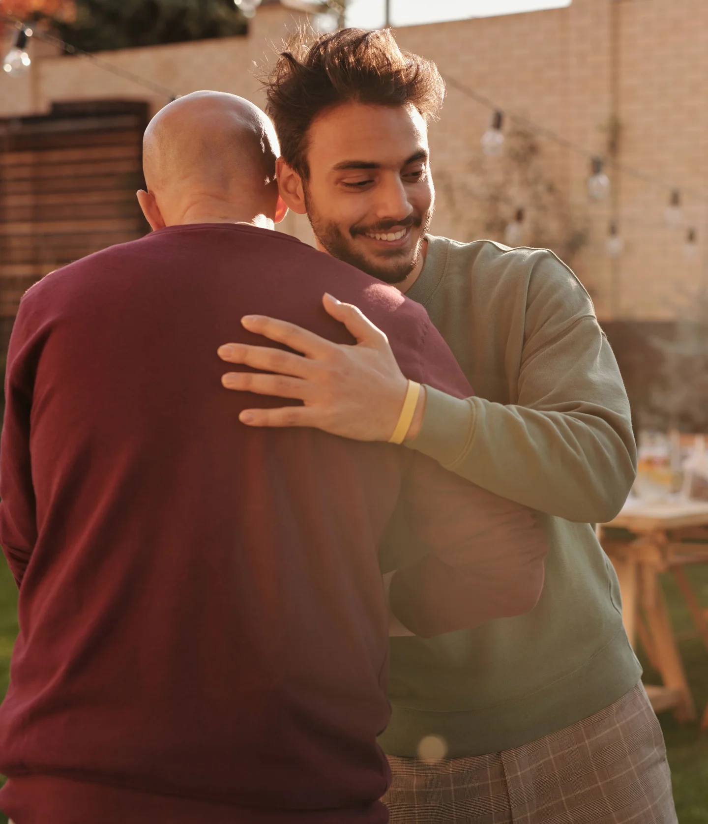 A smiling man with messy hair hugs another person outdoors in a sunlit yard. The other person is facing away from the camera and is bald, wearing a maroon sweater. String lights and a table are visible in the background.