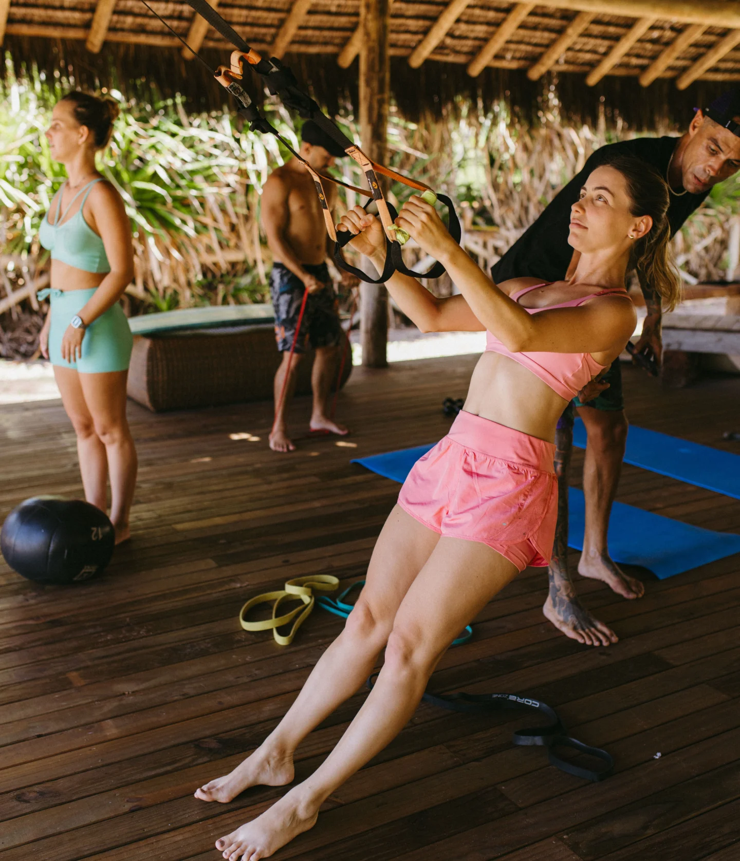 A woman in pink workout clothes uses suspension straps for exercise on a wooden deck, while others in athletic wear stand and observe in the background. The setting appears to be outdoors under a thatched roof.