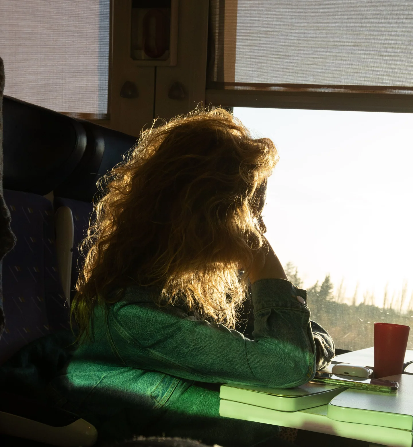 A person with curly hair sits by a sunlit train window, resting their head on their hand and looking outside. A red cup, a phone, and a green notebook are on the table in front of them.