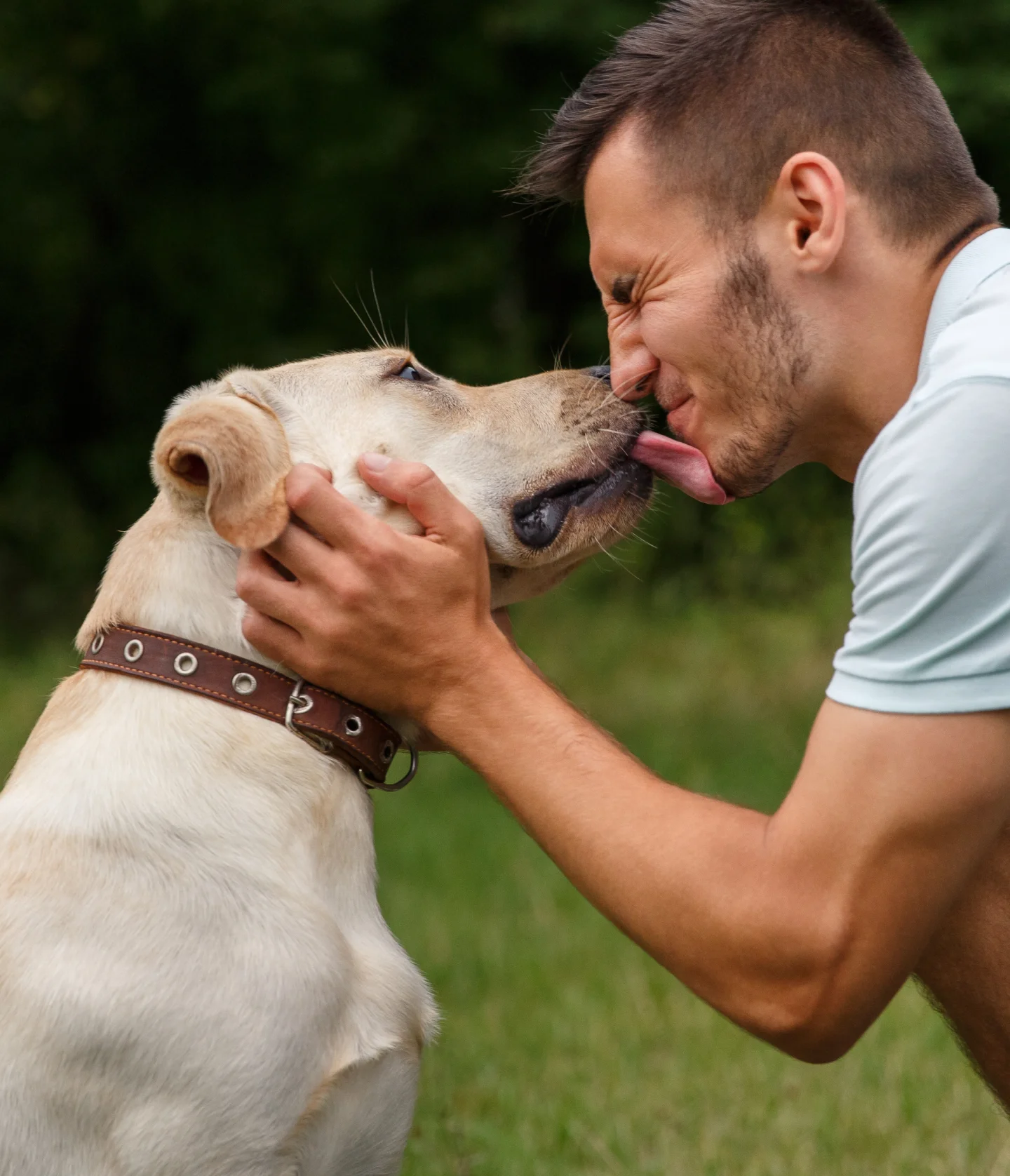 A man kneels on grass, smiling with his eyes closed as a yellow Labrador retriever licks his face. The man gently holds the dogs head, and both appear happy outdoors with greenery in the background.