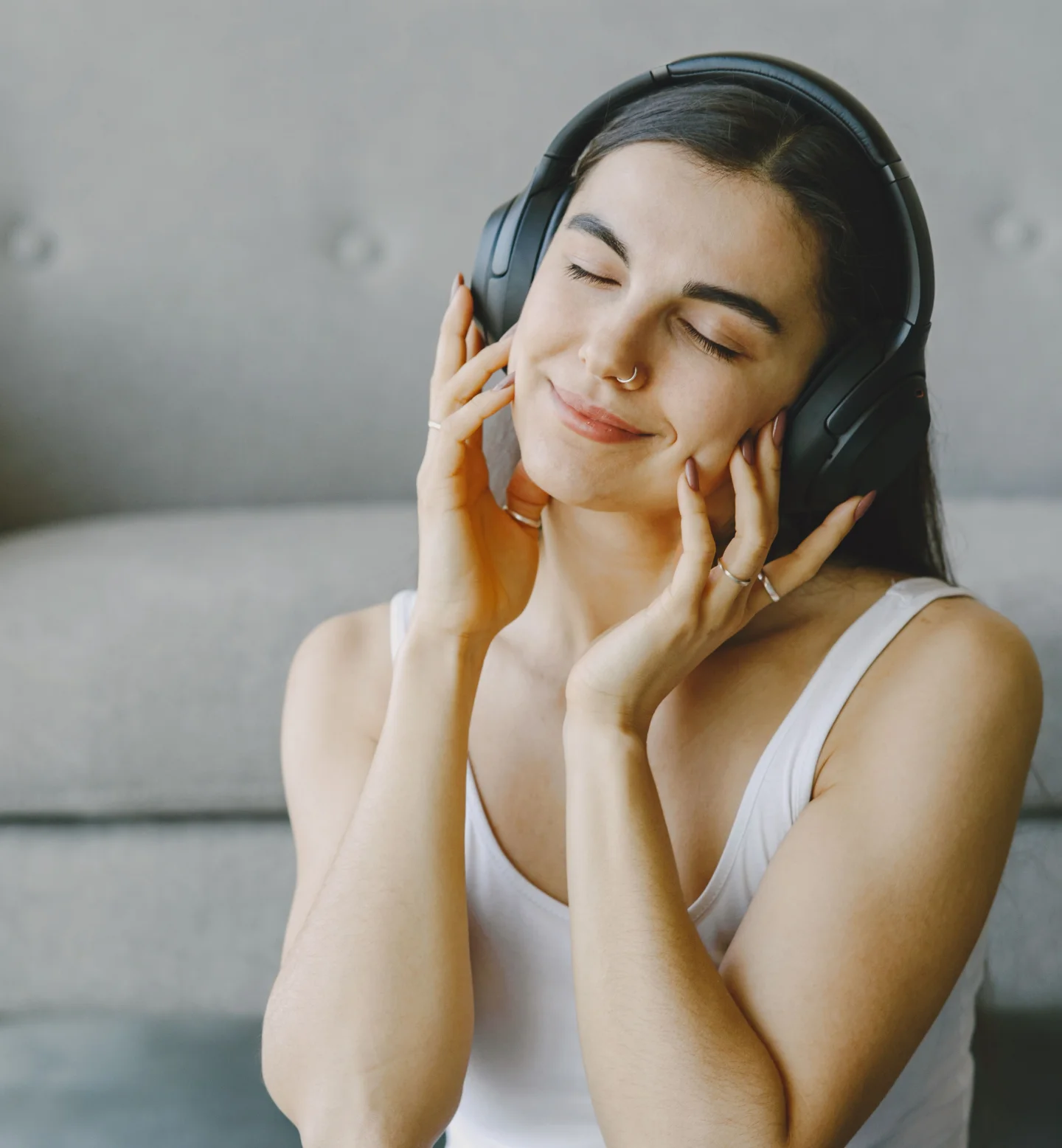 A woman with long dark hair, wearing a white tank top and headphones, sits in front of a sofa with her eyes closed and a peaceful smile, enjoying music.