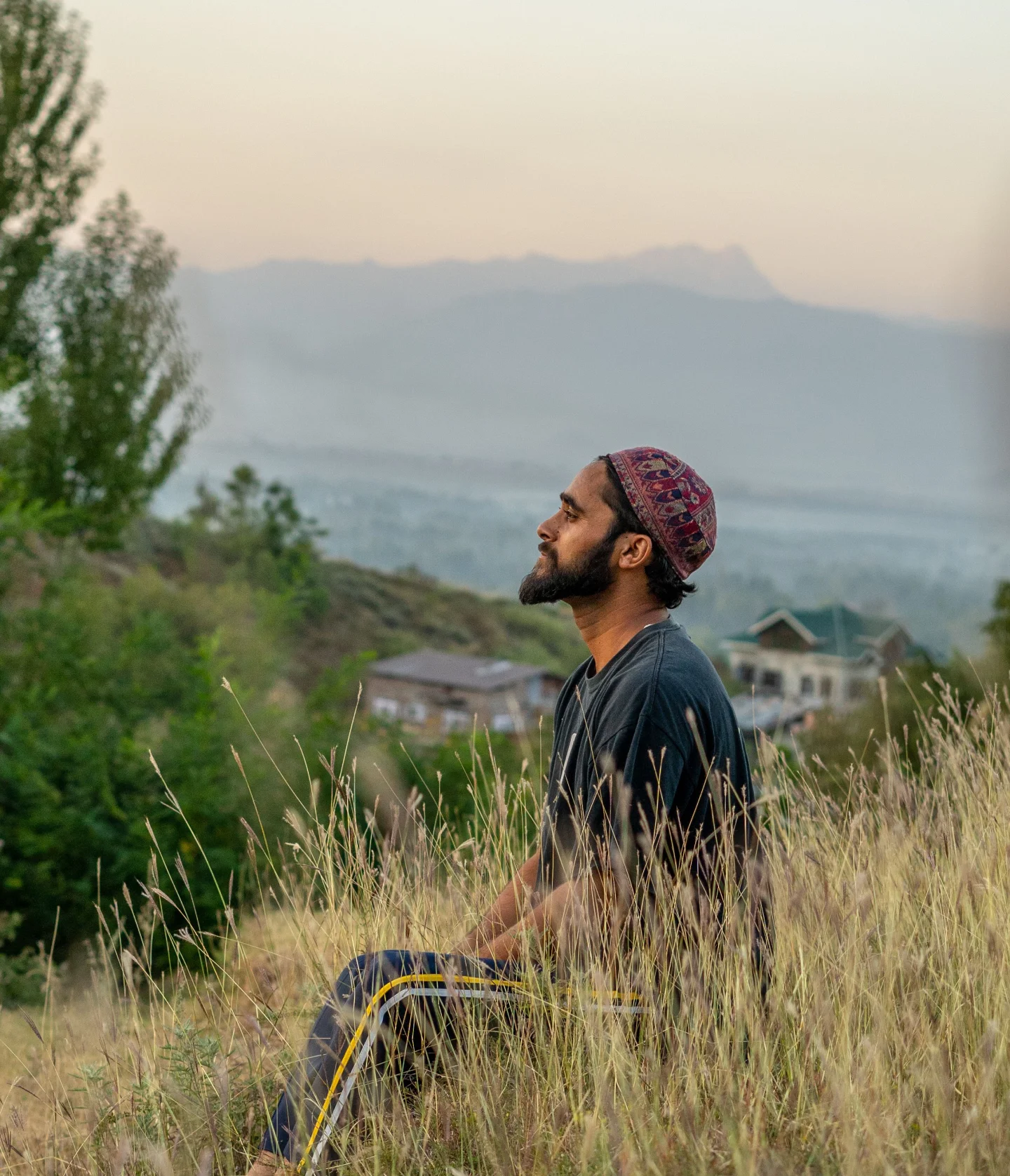 A man with a beard wearing a patterned cap sits in tall grass on a hillside, eyes closed, smiling peacefully. Trees, houses, and distant mountains are in the background under a soft, pastel sky.