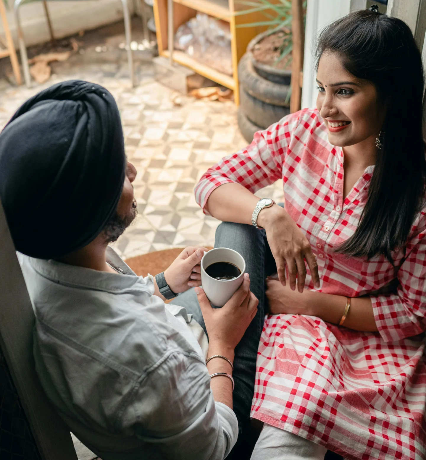 A man and a woman sit closely together, smiling and talking. The man holds a cup of coffee, and the woman wears a red and white checkered dress. They appear relaxed and happy in a cozy indoor setting.