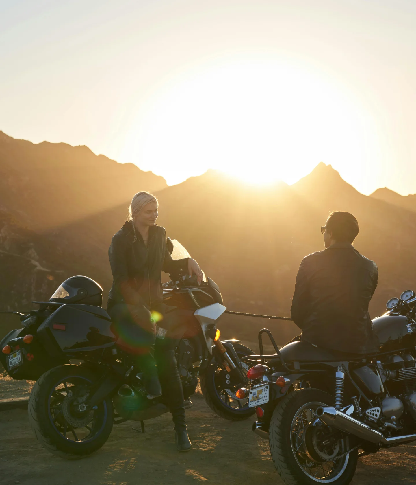 Two people wearing jackets sit on motorcycles, facing each other and talking at sunset, with mountains in the background and warm sunlight filling the scene.