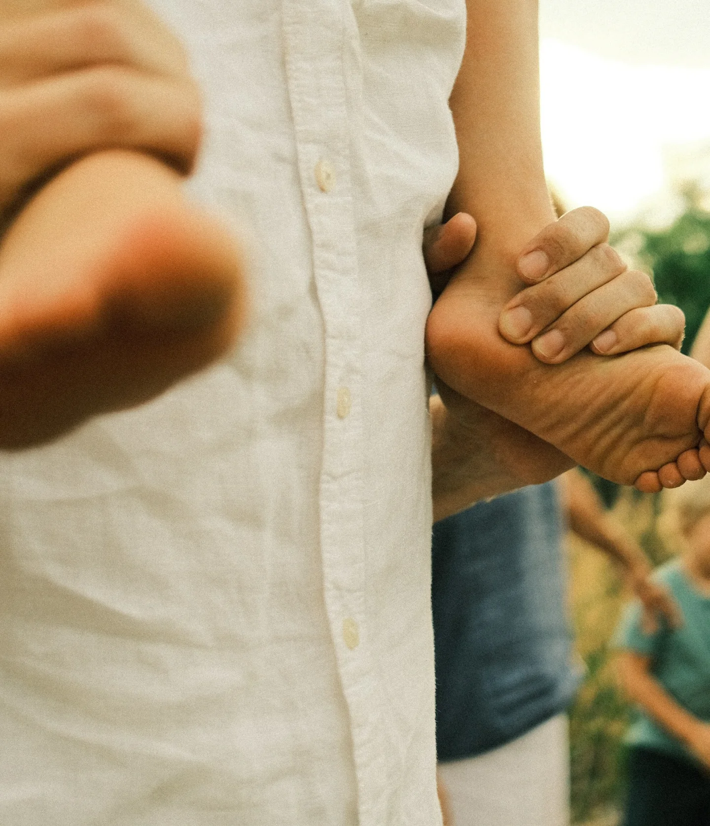 A person in a white shirt carries a barefoot child on their shoulder outdoors, with hands supporting the childs feet. Blurred figures and greenery appear in the background.