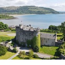 A stone castle with battlements sits by the water, surrounded by green lawns and trees, with hills and a shoreline visible in the background under a partly cloudy sky.