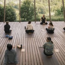 A group of people participate in the Italy Yoga Retreat: Pasta, Hiking & Sunshi, practicing yoga on mats outdoors on a wooden deck amid lush greenery, while an instructor sits at the front facing the group.