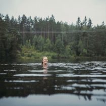 A person smiles while swimming in a lake surrounded by dense green trees under an overcast sky.
