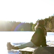 A person in winter clothing sits on a wooden dock by a frozen lake, leaning back and facing the bright sun, with a forest in the background.