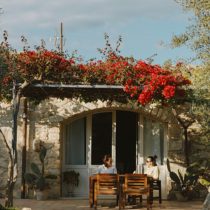 Two people relax at a wooden table outside a stone house with white arched doors during the Sicily Yoga Retreat. Red flowers cascade across the roof, and cacti grow nearby, creating a sunny and peaceful atmosphere perfect for enjoying yoga, pizza, and boat trips.