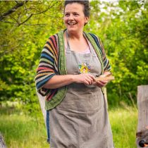 A woman wearing a striped sweater and apron smiles as she holds a small foraged item, standing outdoors in the lush green woods during the Somerset Food & Wellness Retreat: Foraging experience.