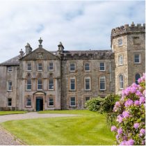A large stone mansion with arched windows, a round turret, and ornate details stands behind a green lawn with blooming pink flowers on the right under a partly cloudy sky.