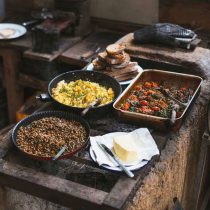 Four rustic dishes from Somerset Food & Wellness Retreat: Foraging are arranged on a stone counter—sautéed greens, scrambled eggs with herbs, roasted vegetables, and a slab of butter with a knife—while toasted bread is stacked behind them.