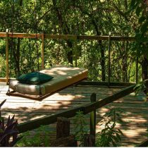 A single outdoor hanging bed with a green pillow sits on a wooden deck surrounded by lush green trees in sunlight.
