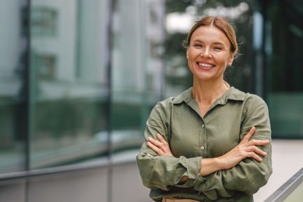 A woman stands outdoors, smiling with arms crossed, wearing a green shirt and beige high-waisted pants, next to a glass building and railing—a confident image that captures the spirit of a midlife career change or someone exploring how to switch careers at 40.