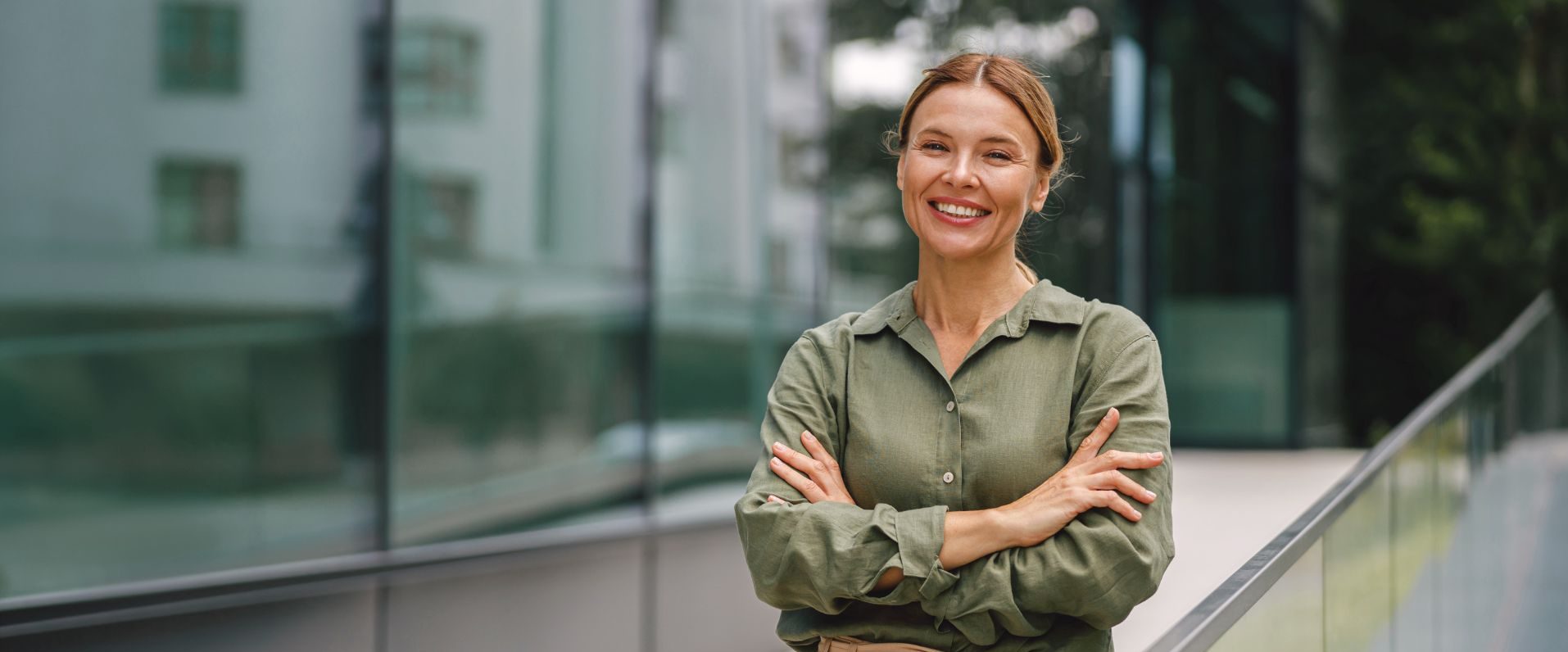 A woman stands outdoors, smiling with arms crossed, wearing a green shirt and beige high-waisted pants, next to a glass building and railing—a confident image that captures the spirit of a midlife career change or someone exploring how to switch careers at 40.