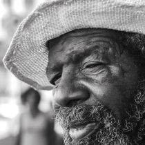 Black and white close-up of an older man with a beard and textured hat, gazing thoughtfully. The background is blurred with a person walking. Text reads zizo bongekile manona art in the bottom left corner.