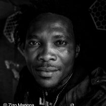 Black and white close-up portrait of a man with short curly hair, looking directly at the camera, wearing a jacket. The name © Zizo Manona appears in the bottom left corner.