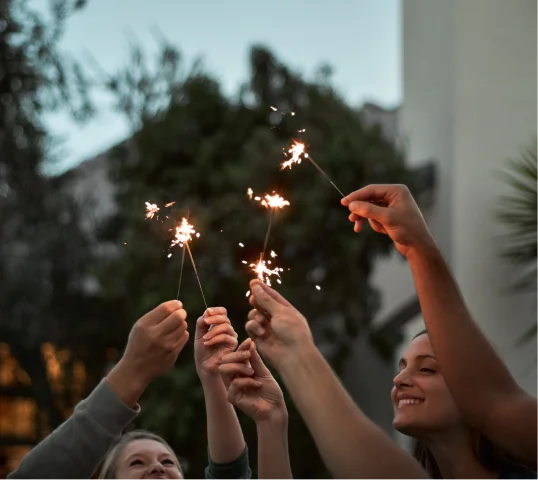 A group of people hold up sparklers together outdoors at dusk, smiling and celebrating personal growth, with trees and a building blurred in the background.