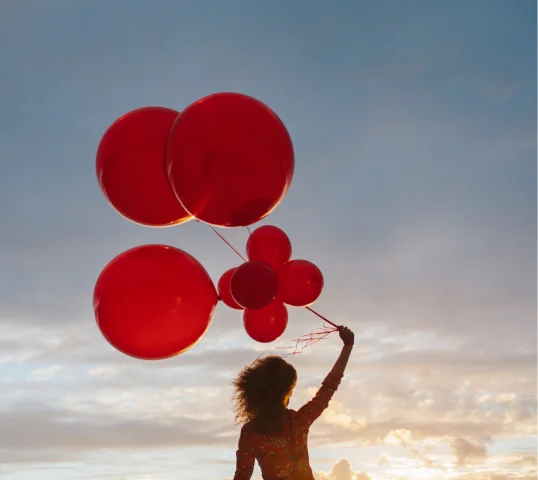 A person stands outdoors at sunset, holding a bunch of large red balloons against a cloudy sky—a quiet moment that reflects personal growth and renewal.