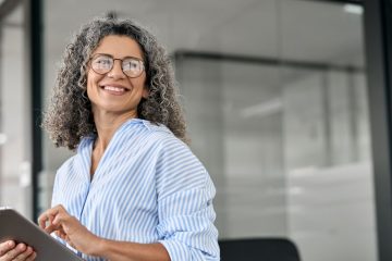 Confident mature professional woman with curly gray hair holding a tablet in a modern office