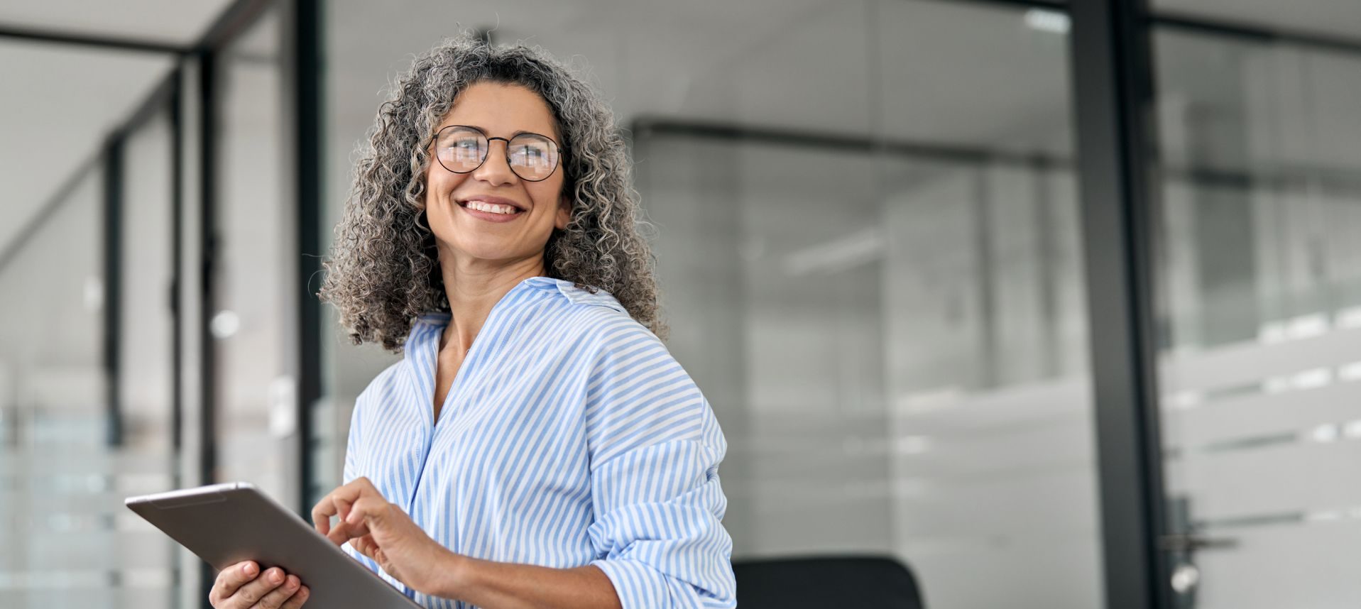 Confident mature professional woman with curly gray hair holding a tablet in a modern office